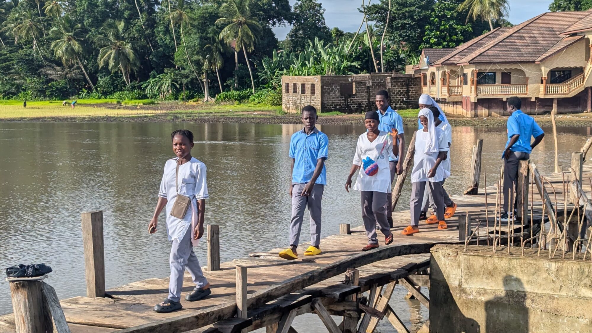 School pupils carefully walk across a locally made wooden bridge — an unstable, short term structure that often collapses within months due to strong river water flow and wood rot